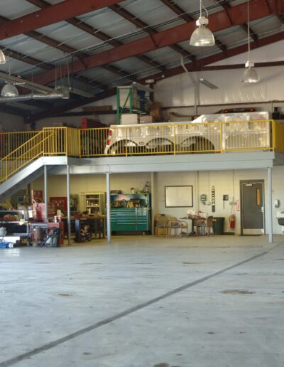 Industrial warehouse interior with a mezzanine, workstations, and scattered equipment.