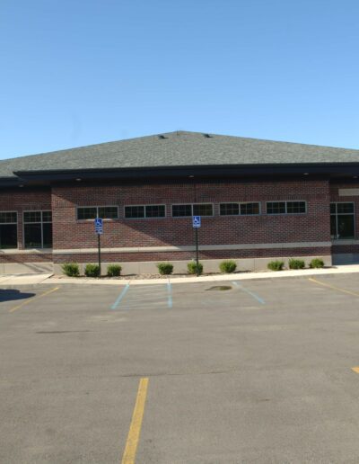 A single-story brick building with a black roof and a parking lot in the foreground under a clear blue sky.